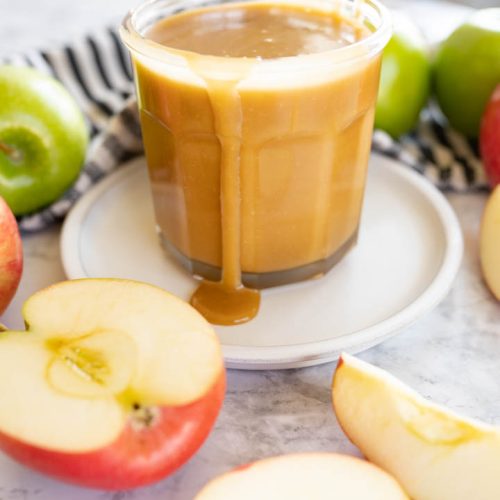 A cup of caramel sauce sitting on the counter with apple slices around it.