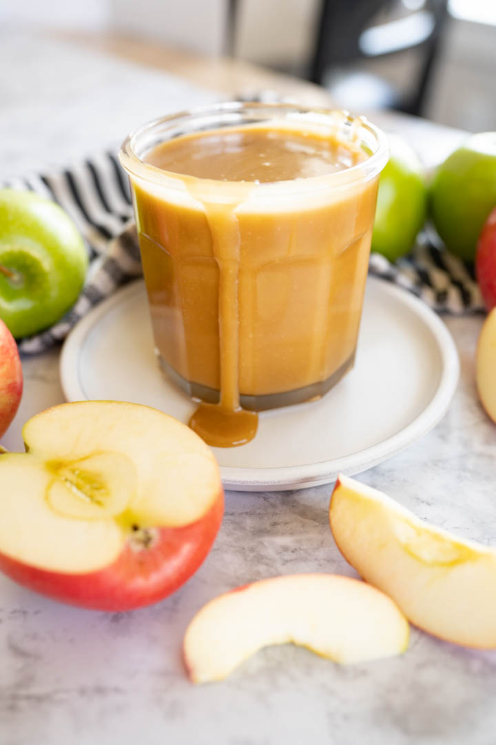 A cup of caramel sauce sitting on the counter with apple slices around it.