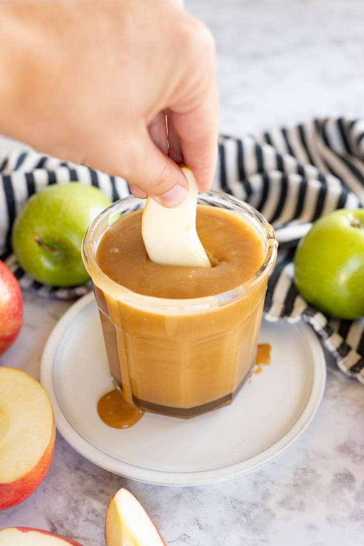A hand dipping an apple slice into a jar of caramel sauce. 