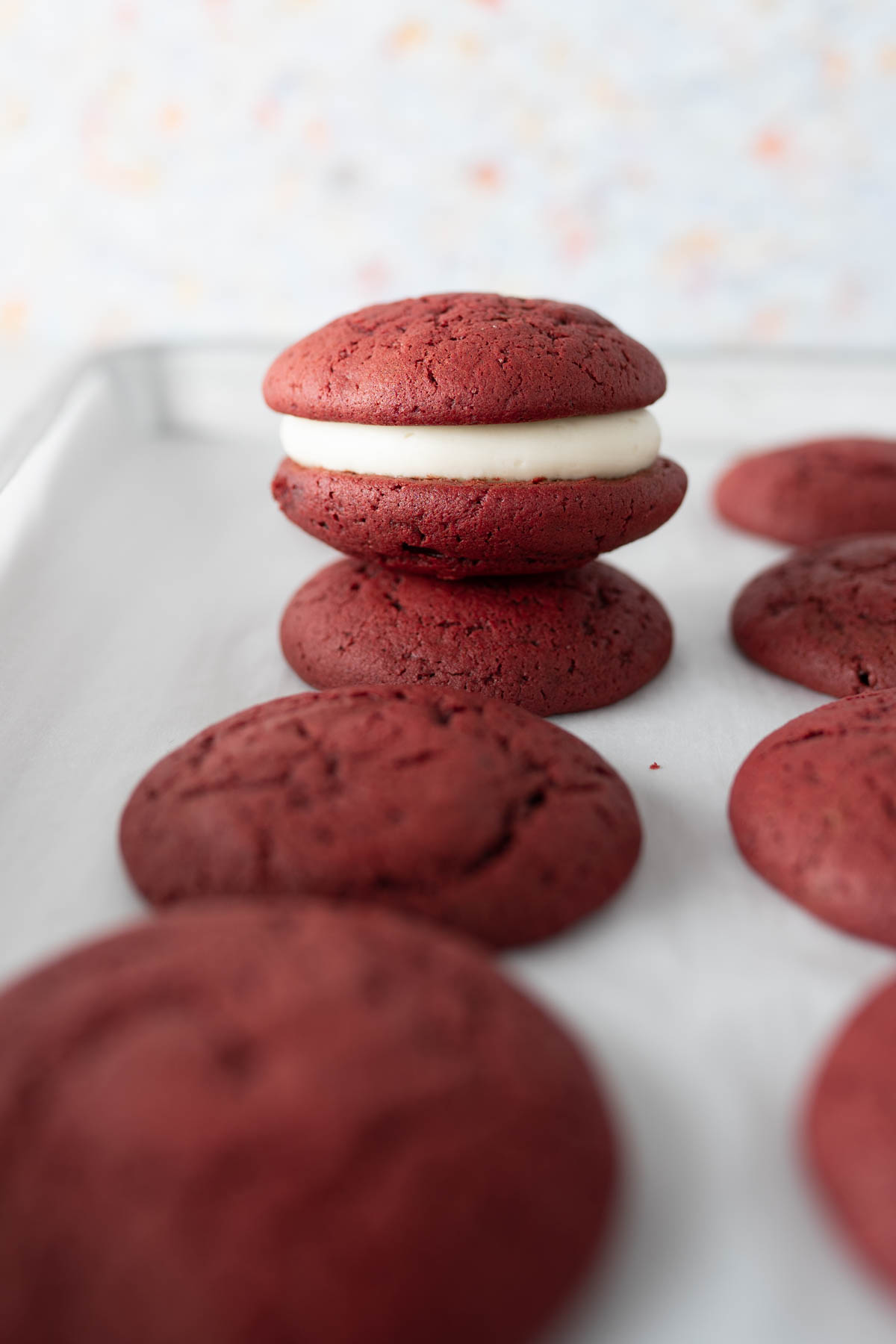 A completed red velvet whoopie pie stacked on top of the red velvet cookies on a cookie sheet. 