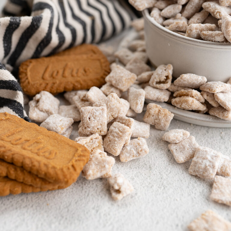Cookie butter puppy chow spilling out onto the counter from a bowl with biscoff cookies.